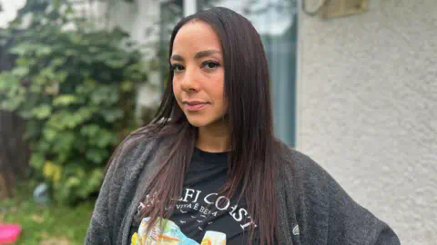 A young woman with long dark hair stands in a garden, wearing a grey cardigan and a t-shirt featuring a painting of the Amalfi Coast in Italy. 