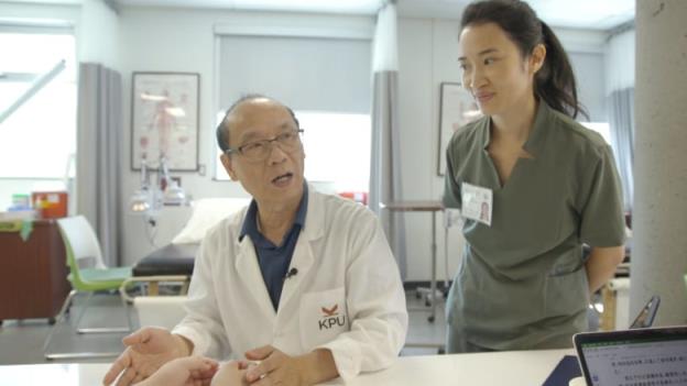 A man in a white coat with the acronym KPU sits at a table, holding a volunteer's hands as he speaks to students in a medical lab setting. A student stands to his right.