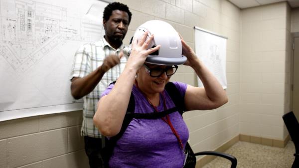 Clack stands behind Chaidez with a hand to the back of the helmet, which resembles a white hard hat, as Chaidez holds it at the sides.